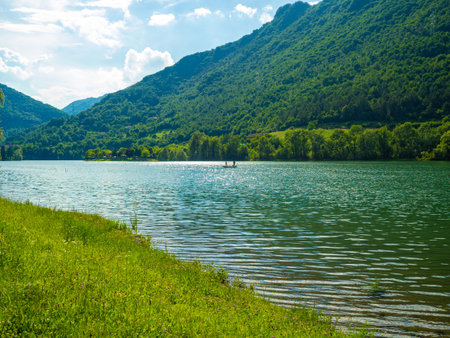 Stunning landscape featuring a calm lake reflecting the cloudy sky. Majestic mountains rise in the distance, framed by lush greenery, creating a tranquil and picturesque scene. Lake Idro, Crone, Italyの写真素材
