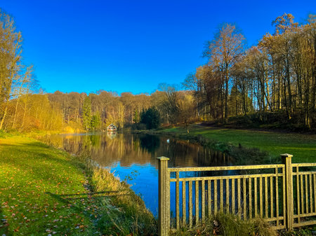 A picturesque scene in waterloo, belgium featuring a serene lake surrounded by lush greenery and an old wooden fence. The vibrant autumn leaves create a charming atmosphere under a clear blue sky.の写真素材