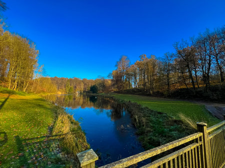 A picturesque scene in waterloo, belgium featuring a serene lake surrounded by lush greenery and an old wooden fence. The vibrant autumn leaves create a charming atmosphere under a clear blue sky.の写真素材