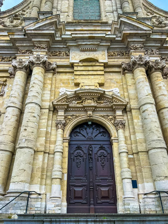 This image captures a striking front view of a baroque church in Leuven, showcasing its intricate stone facade, majestic stairs leading to the front door, and a vibrant blue sky.の写真素材
