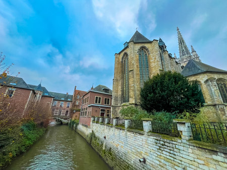 A picturesque view of a gothic church juxtaposed with historic townhouses in Leuven, complemented by a serene canal flowing beneath a clear blue sky, showcasing the citys rich architectural heritage.の写真素材