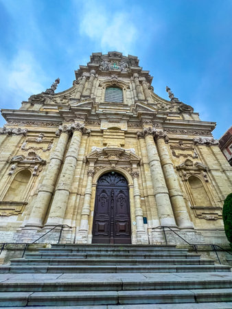 This image captures a striking front view of a baroque church in Leuven, showcasing its intricate stone facade, majestic stairs leading to the front door, and a vibrant blue sky.の写真素材