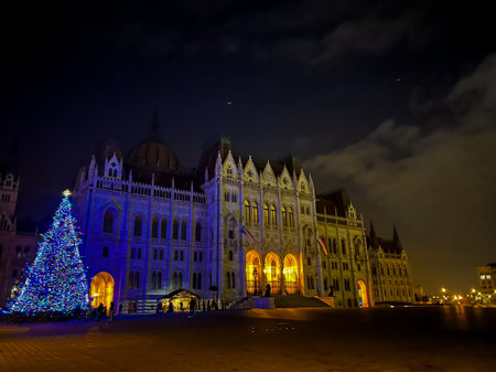 A beautiful night scene showcasing the hungarian parliament building in Budapest, illuminated with vibrant blue lights for christmas, along with a festive christmas tree in the foreground.の写真素材