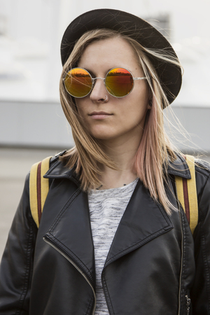 Portrait of young woman in hipster glasses,pot hat and black leather jacket standing and looking forward in cityの写真素材