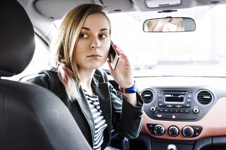 Beautiful young attractive woman sitting in driver seat in the car looking back and holding mobile...
