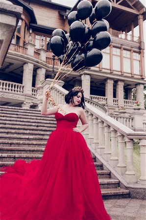 Beautiful fashion young woman in gorgeous red dress and crown sitting holding black balloonsの写真素材