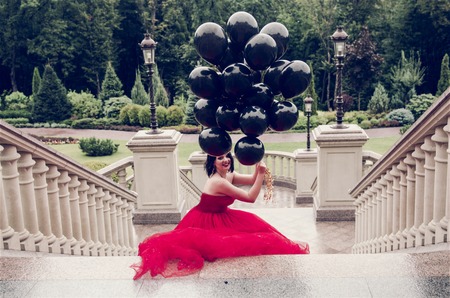 Beautiful fashion young woman in gorgeous red dress and crown sitting holding black balloonsの写真素材