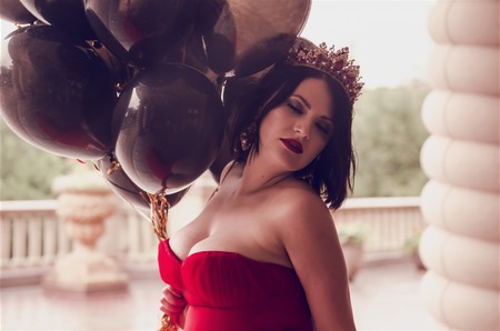 Beautiful fashion young woman in gorgeous red dress and crown sitting holding black balloonsの写真素材