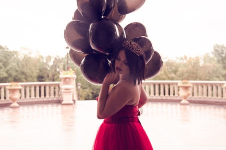 Beautiful fashion young woman in gorgeous red dress and crown sitting holding black balloonsの写真素材