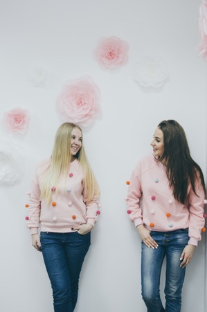 two beautiful young girls posing in the same sweaters against wall with flowersの写真素材