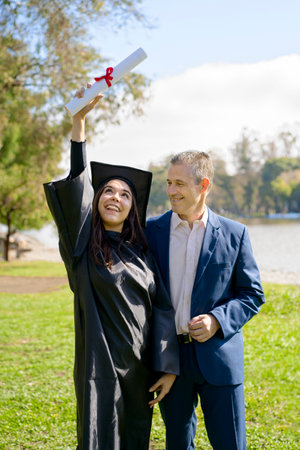 Young girl recently graduated, dressed in cap and gown, with her degree in her hand, celebrating with her father on the university campus. Very happy expression, achievement, beginning of a new stageの写真素材