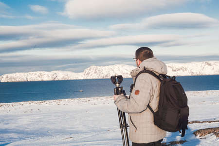 North sea coast with ice and snow. Man blogger at Europe travel adventure trek. Person wearing hat and jacket for cold weather backpack with tripod and photo camera. Horizontal scenic photoの写真素材