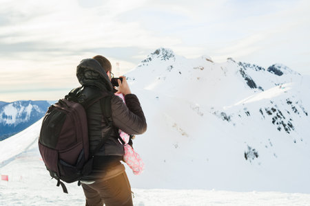 Father with baby child in carrier backpack on snowy slope. Family walking at winter time. Man photographer blogger vlogger traveling with infant outdoor. Healthy vacations in mountains. Copy spaceの写真素材
