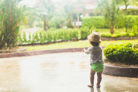 Cute baby toddler girl standing outdoors under the rain with bare feet. Kid is enjoying the warm summer rain. Childhood, freedom, happiness, child day concept. Scenic photo with copy spaceの写真素材