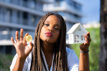 Young latin girl posing with a bitcoin coin and a miniature house.Young investor concept.の写真素材