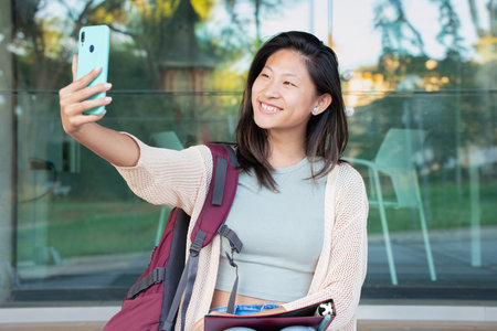 Young Asian woman having a video call with cell phone and smiling outdoors.の写真素材