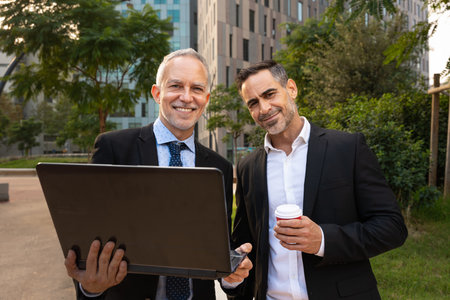 Two businessmen with laptop in hand smiling looking at camera outdoors.