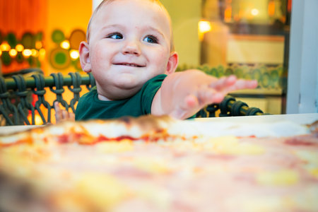Portrait of a smiling lovely boy sitting on a chair wanting to grab pizza from the table.の写真素材