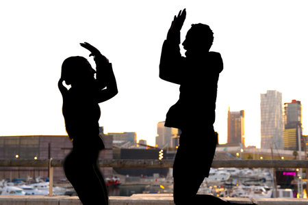 Silhouette of two friends with hands raised at dusk or sunrise outdoors.の写真素材