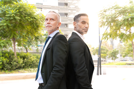 portrait two successful businessmen standing back to back looking at camera.