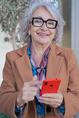 Senior woman looking at camera with a smile, holding a red smartphone outdoors.の写真素材