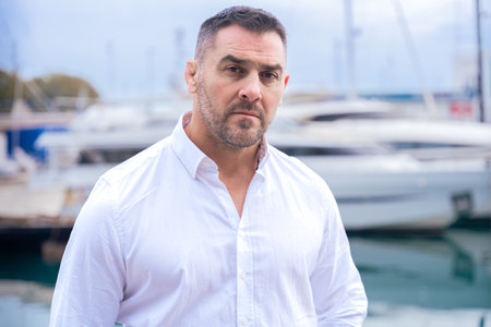 Serious Caucasian man with short dark hair and beard standing near luxury yachts.