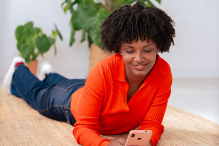 Happy African American woman using smartphone while lying on the floor at home.