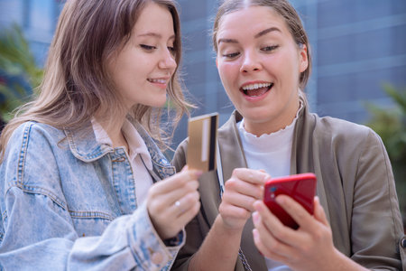Two happy Caucasian women shopping online with a credit card and smartphone outdoorsの写真素材
