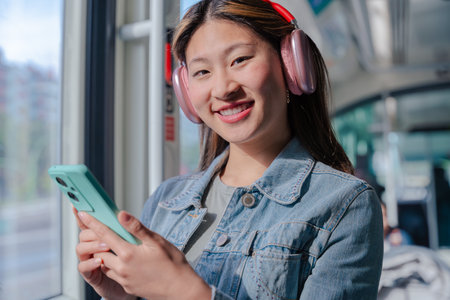 Smiling Chinese woman with headphones using smartphone while riding city tram looking at camera