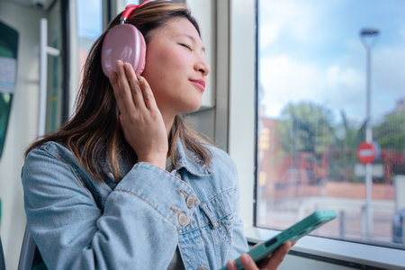 Chinese woman enjoying music with headphones while riding tram and holding phoneの写真素材