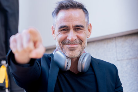 Smiling businessman with headphones pointing at camera while standing outdoors in styleの写真素材