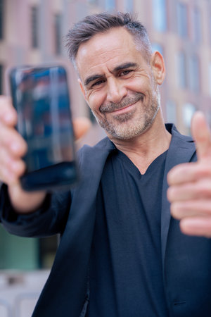 Smiling businessman showing smartphone and giving thumbs up in city setting looking at cameraの写真素材