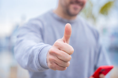 Unrecognizable Caucasian man smiling and giving thumbs up while holding smartphone outdoors