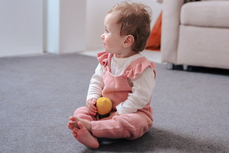 Cute baby girl sitting barefoot on carpet holding toy in bright cozy home interior