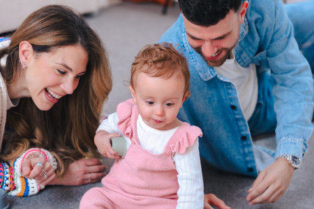 Parents play on floor with baby girl in pink overalls holding toy at home