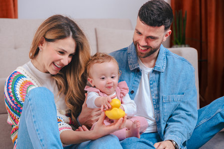 Parents show baby girl plastic duck toy while relaxing together on sofa at home
