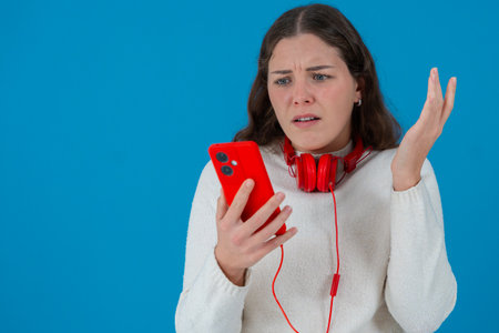 Worried young woman reacts in shock while looking at smartphone with red headphones