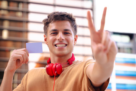 Smiling young man with headphones showing blank card and making peace sign outdoorsの写真素材