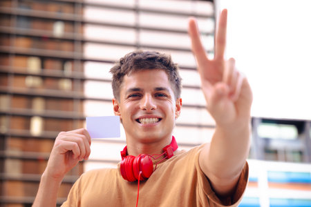Smiling young man with headphones showing blank card and making peace sign outdoorsの写真素材