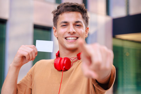 Smiling young man pointing at camera and holding blank business card outdoorsの写真素材