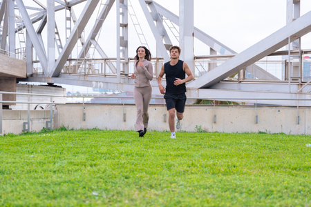 Young man and woman jogging together outdoors green grass under a large bridge