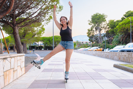 Happy young woman roller skating outdoors in summer, arms raised