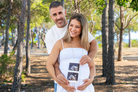 Pregnant couple hugging and holding ultrasound photo outdoors in green parkの写真素材