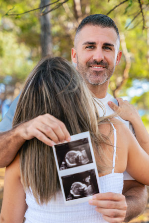 Smiling father looking at camera while pregnant wife holds ultrasound photoの写真素材