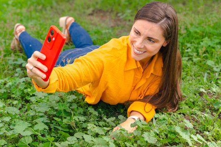 Smiling woman in yellow shirt taking selfie with red smartphone, lying on grass.