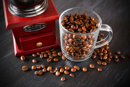 Retro coffee grinder and glass transparent cup full of brown coffee beans and grains around on dark surface. Close-upの写真素材