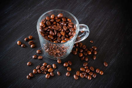 Transparent cup full of brown coffee beans and grains around on dark surfaceの写真素材