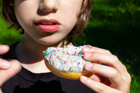 Child eating white glazed donut holding it in his hand outdoor. Part of the face.