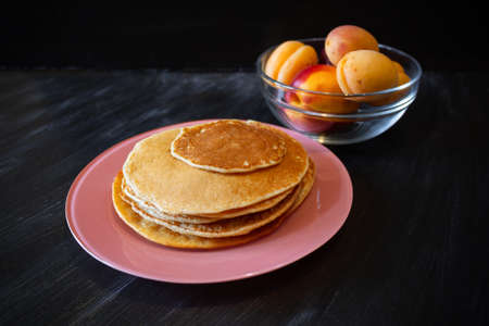 Pancakes on pink plate, nectarines and apricots in transparent bowl on dark surface.の写真素材