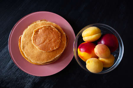 Pancakes on pink plate, nectarines and apricots in transparent bowl on dark surface, view from above.の写真素材